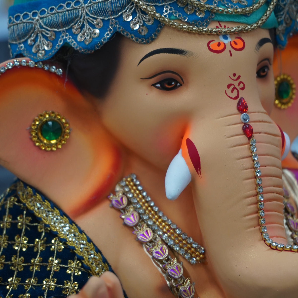 A close-up of a decorated Lord Ganesha idol with a turquoise crown, gold jewelry, and a dark blue patterned garment, shown against a softly blurred background.