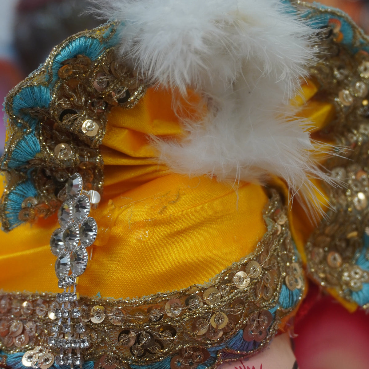 Close-up of a decorated Ganesha idol or figurine, showing the yellow turban wih golden sequin work, white feather and teal.
