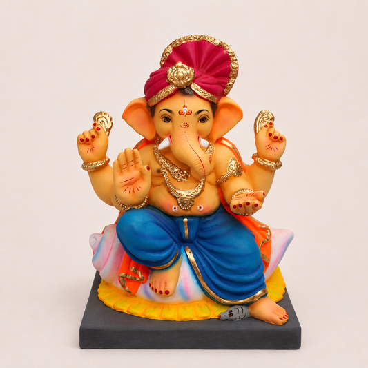 A colorful statue of Lord Ganesha seated on a black rectangular base, wearing a magenta turban with gold trim, blue pants, and gold jewelry, photographed against a plain light beige background.
