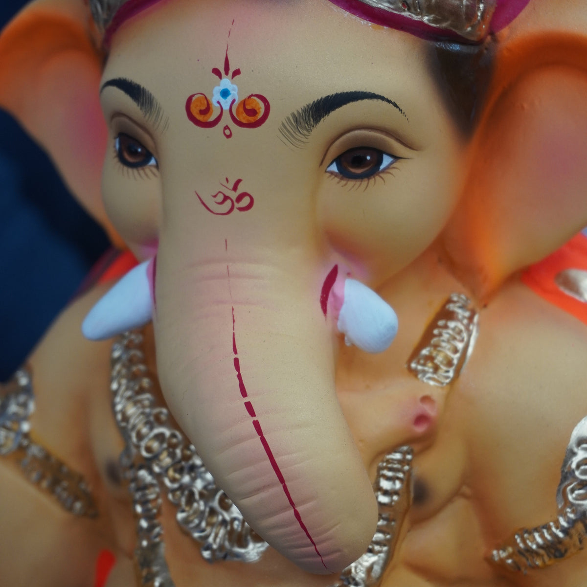A close-up of a Lord Ganesha idol with a pink turban, silver ornaments, and a beige face and trunk, shown against a dark background.