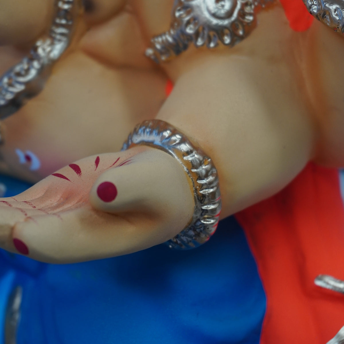 Close-up of a lord ganesh ji with a beige skin-toned body, wearing bright orange and blue garments and multiple ornate silver arm and wrist ornaments.
