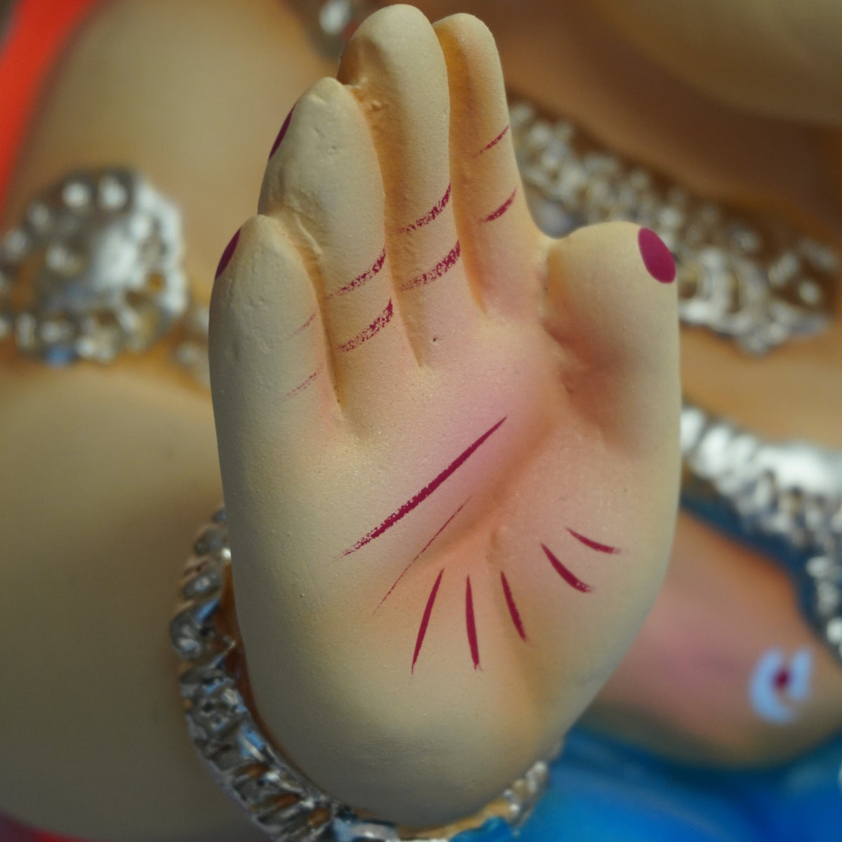 Close-up of a decorative hand sculpture with painted red markings on the palm and fingers, wearing a silver chain bracelet, against a softly blurred multicolored background.