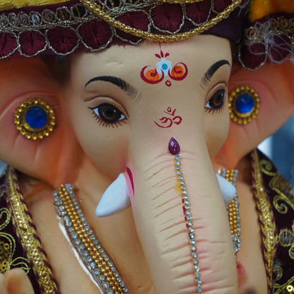 Close-up of a decorative Lord Ganesha idol with a painted elephant head, ornate gold jewelry, blue gem accents, and embroidered maroon and yellow fabric details. 