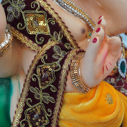 Close-up of a ganesha hands, wearing ornate gold jewelry, a dark maroon embroidered sash, and a bright yellow draped cloth, with green fabric visible at the bottom.