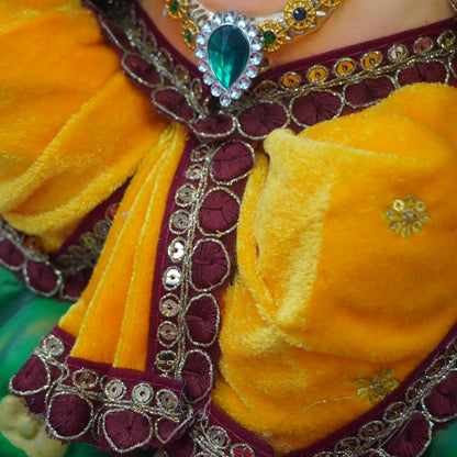 Close-up of a traditional Indian doll or deity figurine wearing a bright yellow and maroon embroidered outfit with ornate gold jewelry and a large green teardrop pendant necklace.