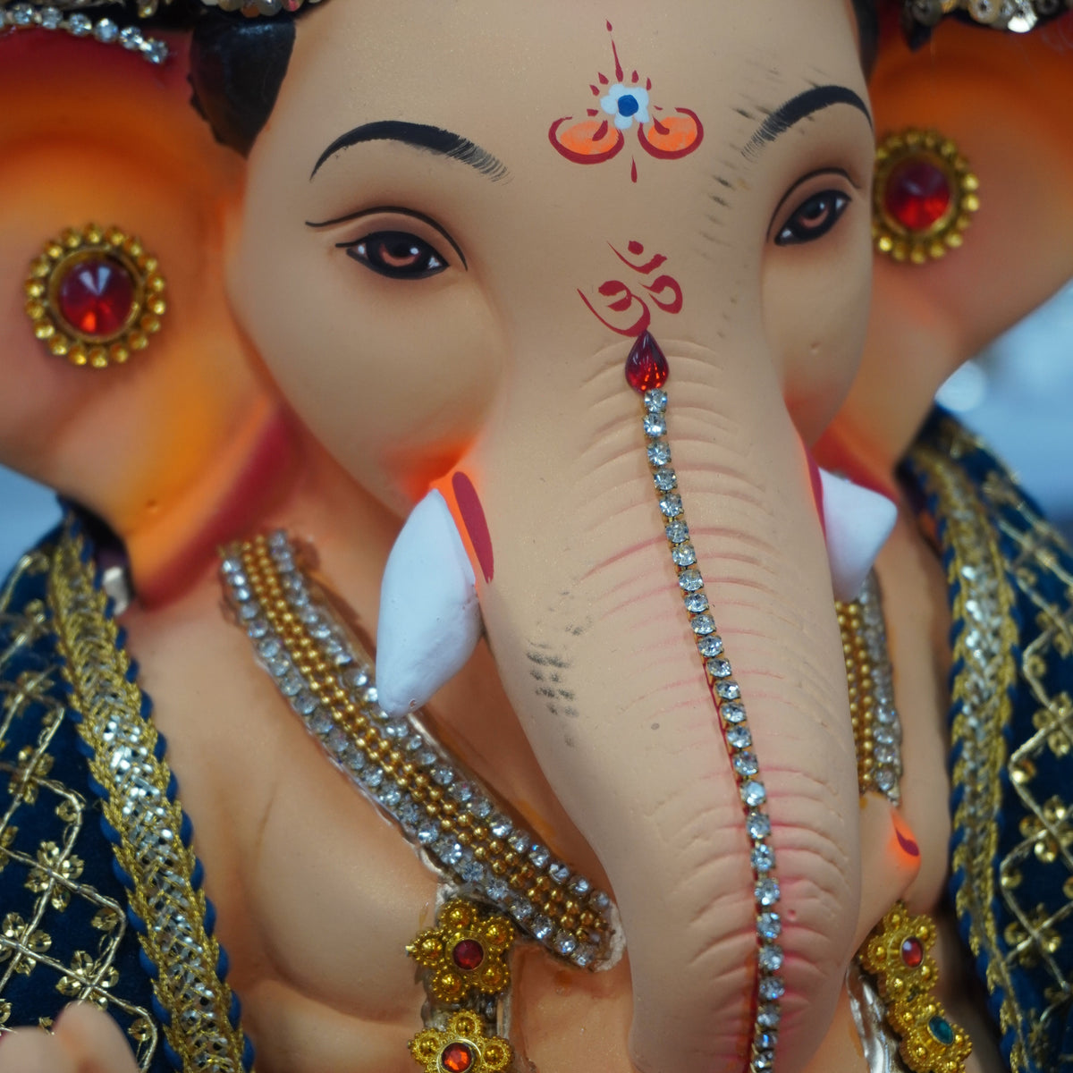 A close-up of a decorative Ganesha idol with a beige elephant head, ornate gold jewelry, a purple crown, and red and blue painted details on a blurred background.