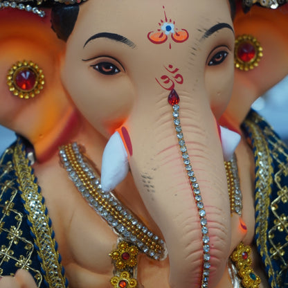 A close-up of a decorative Ganesha idol with a beige elephant head, ornate gold jewelry, a purple crown, and red and blue painted details on a blurred background.
