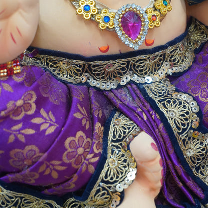 Close-up of a Ganesha statue wearing a purple sari with gold floral patterns and a large ornate gold necklace with a pink gemstone.
