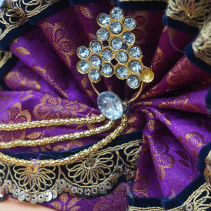 Close-up of a decorative purple and gold fabric headpiece with jeweled embellishments, shown on a doll or figurine face against a neutral background.