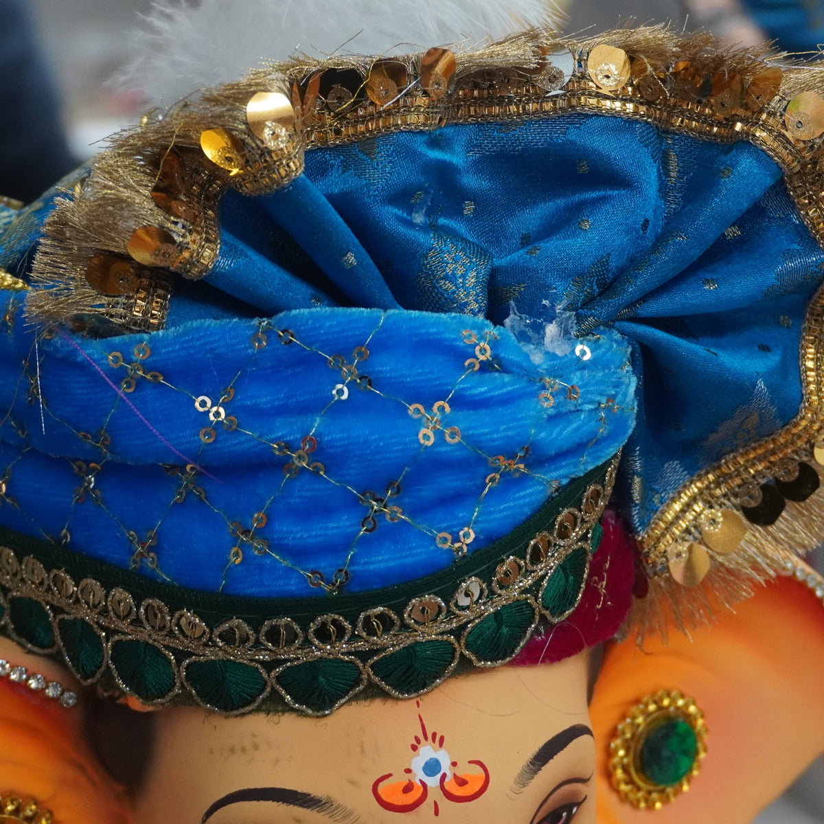 A close-up of a decorated Ganesha idol with a bright blue turban, gold sequins, and ornate jewelry, shown in a real indoor setting with other items blurred in the background.