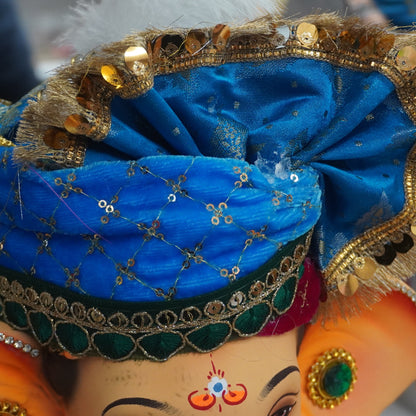 A close-up of a decorated Ganesha idol with a bright blue turban, gold sequins, and ornate jewelry, shown in a real indoor setting with other items blurred in the background.