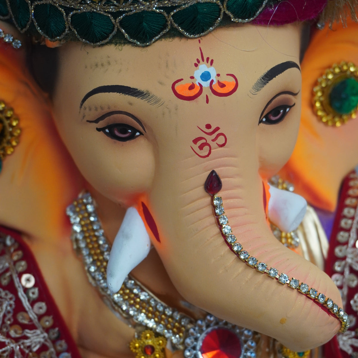 A close-up of a decorated Ganesha idol with a painted elephant head, ornate jeweled headpiece, silver-toned jewelry, and bright orange and red fabric details.