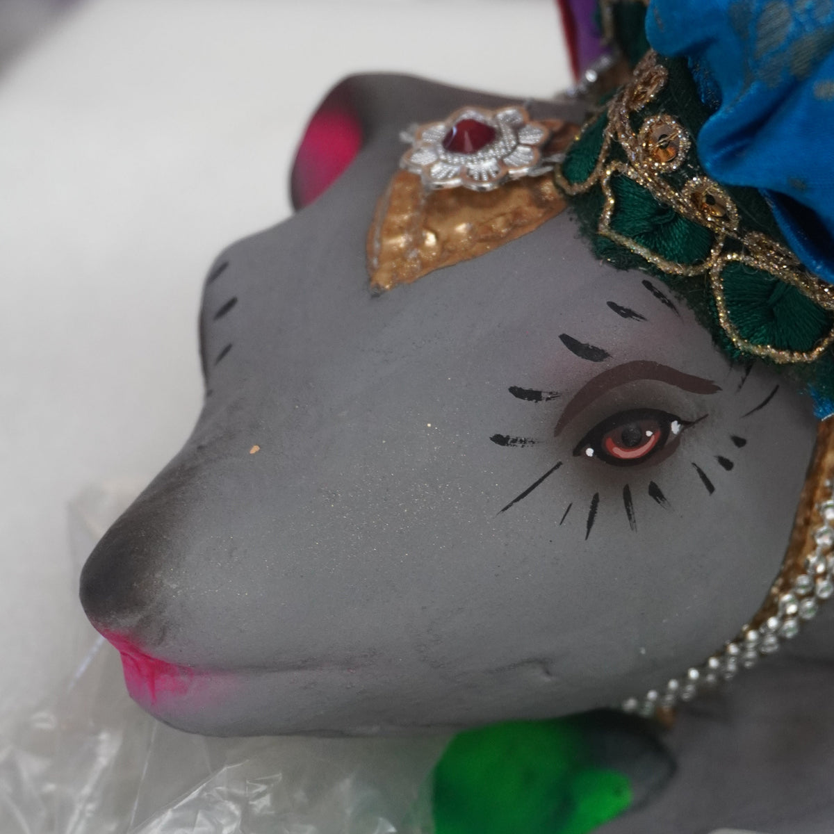 A close-up of a decorative gray animal figurine with painted facial features, a blue embroidered garment, and ornate gold and green headpiece, resting on white foam packaging material.