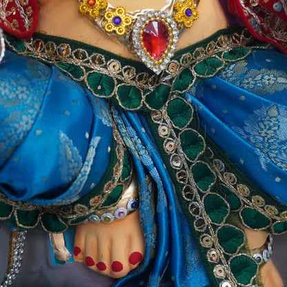 A close-up of a person wearing ornate jewelry, including a silver-toned necklace with red and blue stones, a red and blue embroidered top, and a blue sash with green and silver embellishments.