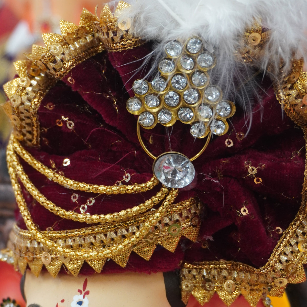 A close-up of a decorated Lord Ganesha idol wearing an ornate maroon velvet and gold turban with a white feather, jeweled headpiece, and blue embroidered clothing.