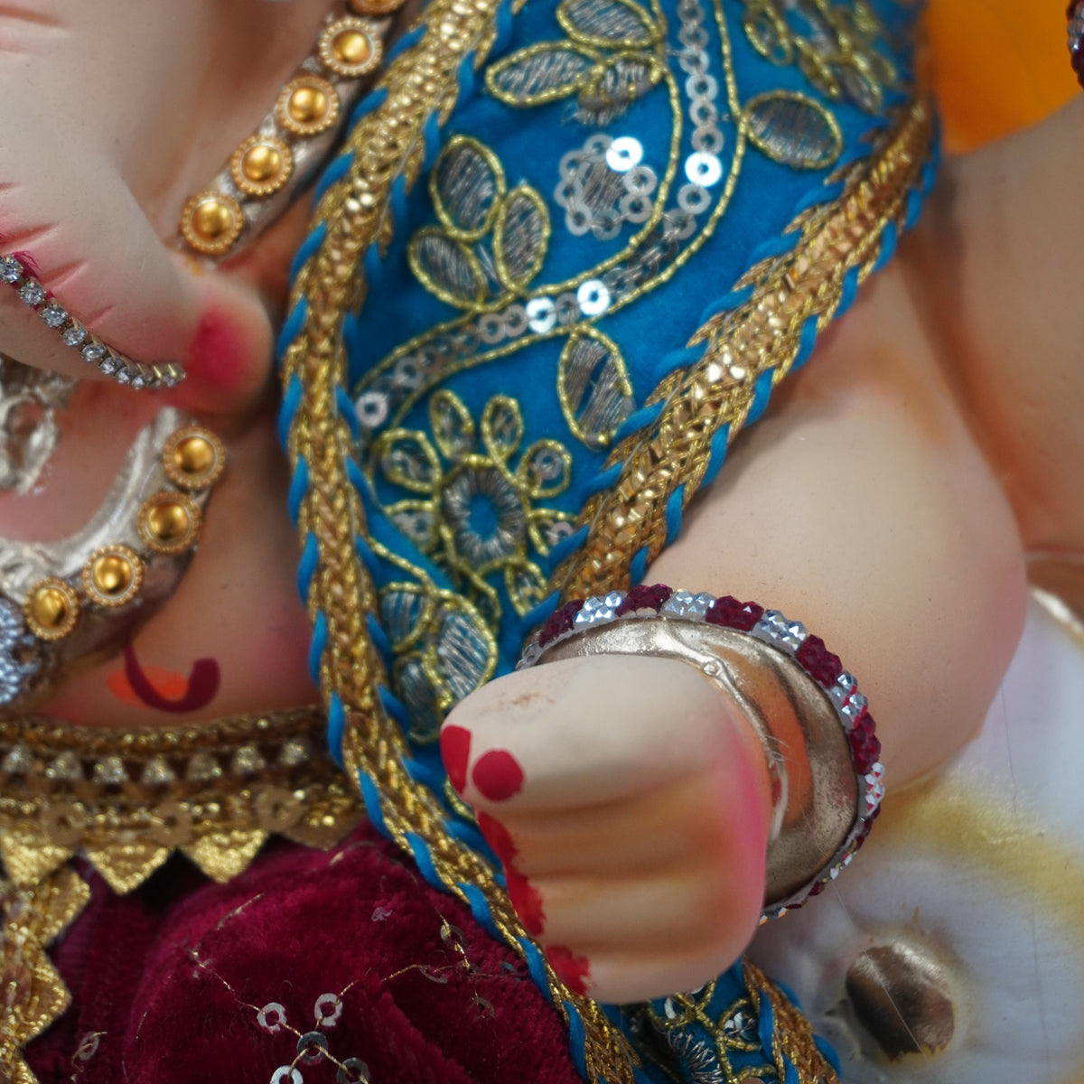 Close-up of a decorated Ganesha idol showing the hand, adorned with silver bangles and a blue sash with gold embroidery, wearing a yellow garment and set against a red fabric background.