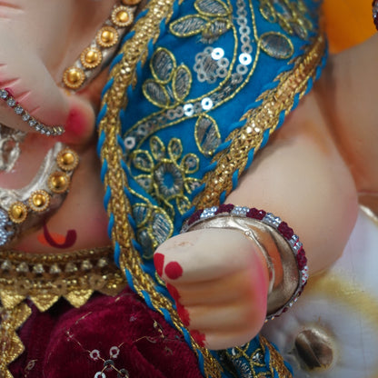 Close-up of a decorated Ganesha idol showing the hand, adorned with silver bangles and a blue sash with gold embroidery, wearing a yellow garment and set against a red fabric background.