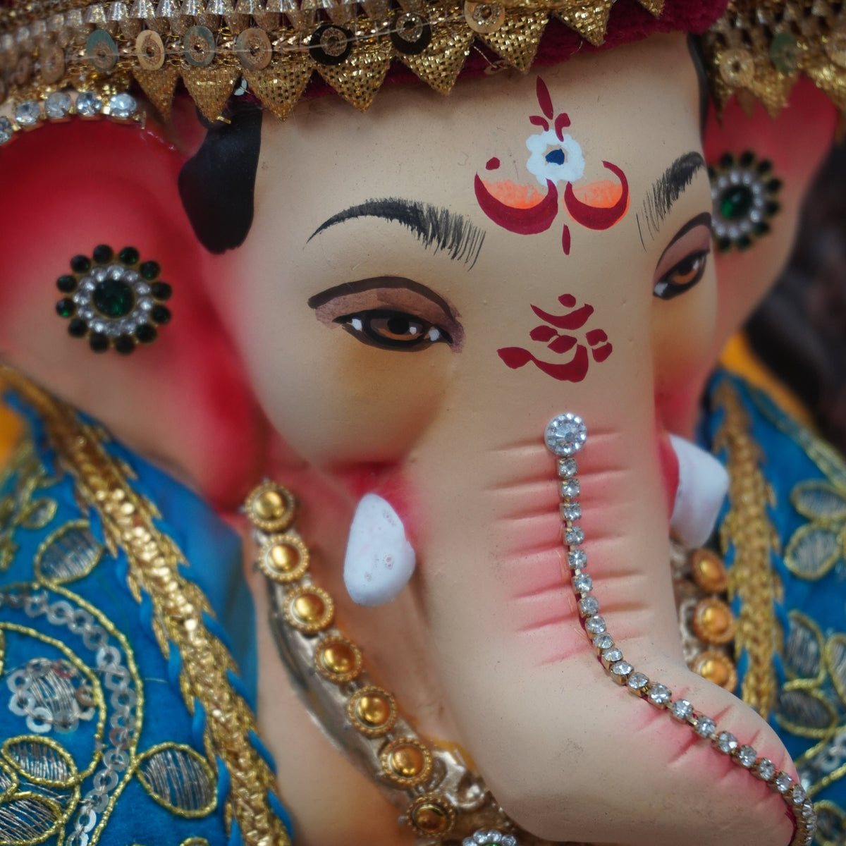 Close-up of a decorated Ganesha idol with a pink face, gold ornaments, a blue embroidered garment, and red forehead markings.