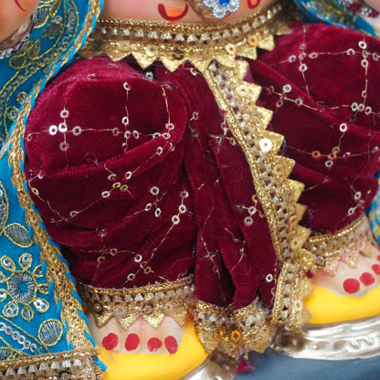Close-up of a decorated Ganesha idol showing the face, blue embroidered drape, gold jewelry, and deep red fabric with sequins.