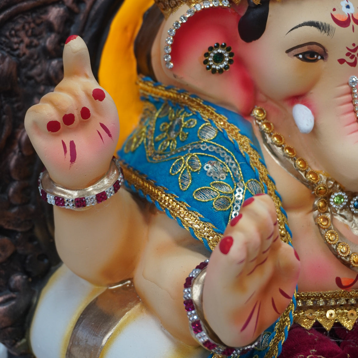 A close-up of a decorated Lord Ganesha statue, showing his face, trunk, and hands with a blue and gold sash, a maroon turban, and red nail polish.