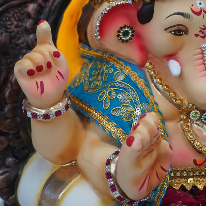 A close-up of a decorated Lord Ganesha statue, showing his face, trunk, and hands with a blue and gold sash, a maroon turban, and red nail polish.