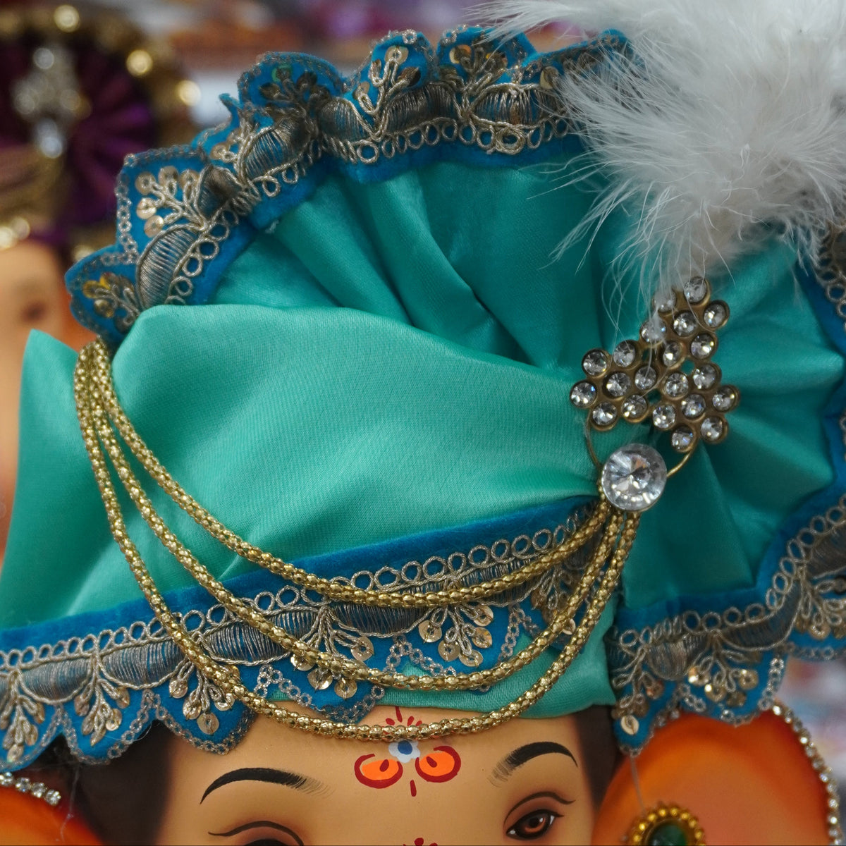 A close-up of a decorated Ganesha idol with a turquoise and blue feathered turban, gold jewelry, and orange ears, shown in a shop setting with other colorful idols blurred in the background.