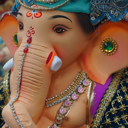 A close-up of a decorated Ganesha statue with a turquoise headpiece, ornate gold jewelry, pink and green gemstone accents, and a blue embroidered garment.
