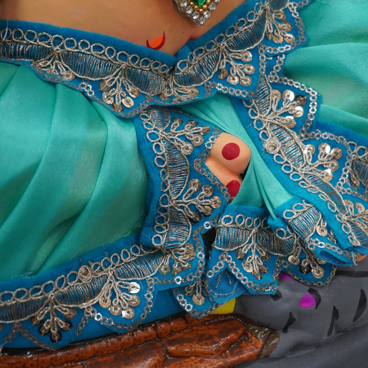 Close-up of a turquoise blue embroidered garment with silver mirror-work borders, a jeweled necklace, and a small peek of a person's hand with red nail polish.