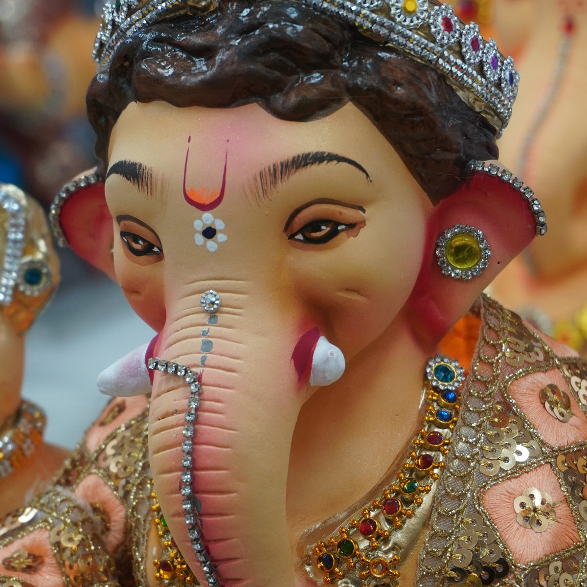 A close-up of a decorative Ganesha idol with a pinkish-beige face and trunk, wearing an ornate jeweled crown and embellished clothing with multicolored stones.