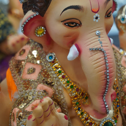 A close-up of a decorative Ganesha idol with a pinkish face, dark hair, white tusks, and ornate gold jewelry with multicolored gemstone accents.