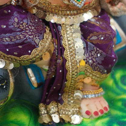 Close-up of a decorated Hindu deity figurine, showing ornate purple and gold clothing with sequins, a large green gem necklace, and a painted foot with red toenails on a green base.