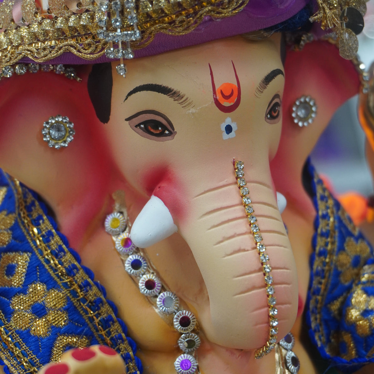 A close-up of a decorative Ganesha idol with a pink face, purple turban, gold embellishments, and blue and gold clothing, shown in high detail.