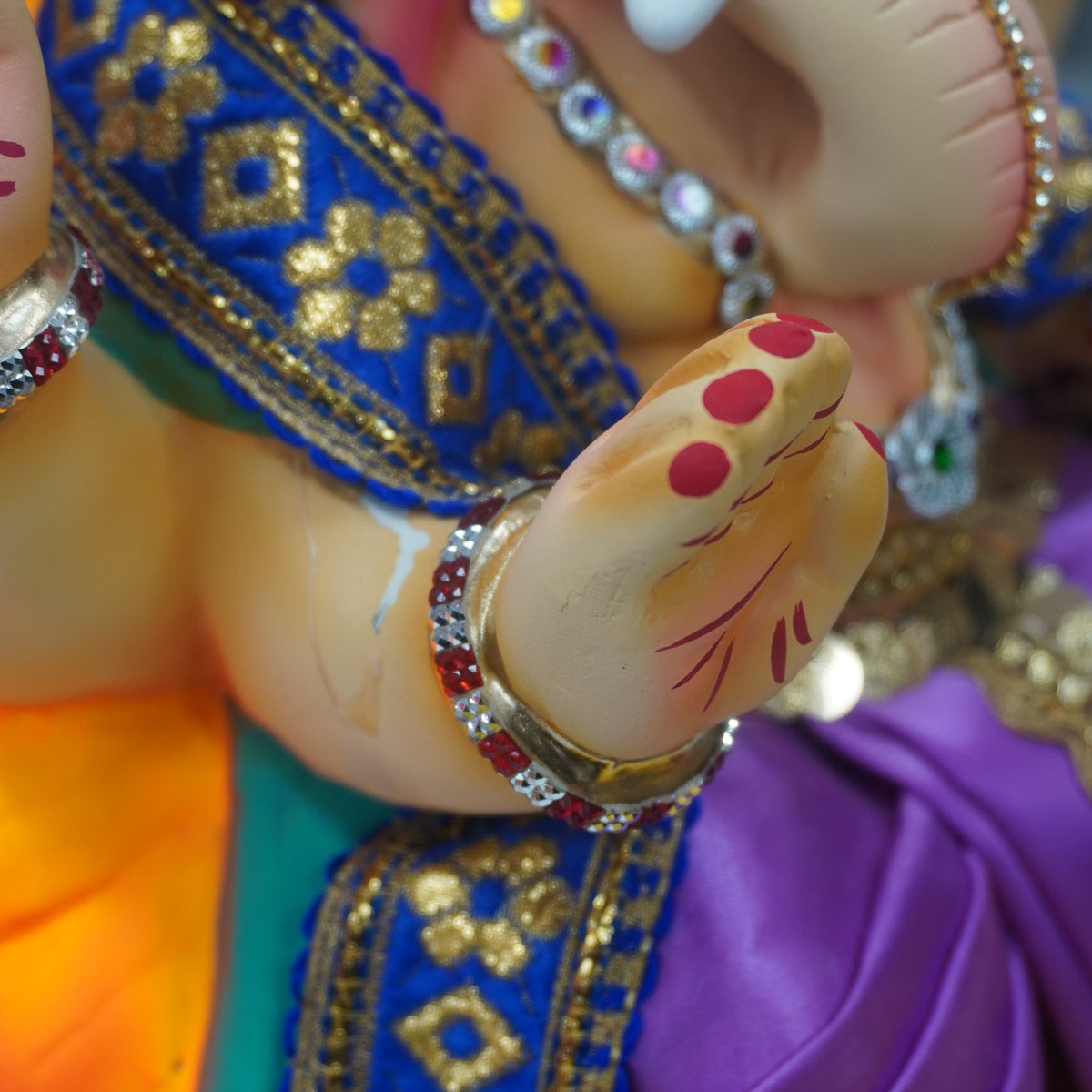 Close-up of a decorative figurine or idol with a beige face and hand, adorned with red markings and jewelry, wearing a blue embroidered sash and purple fabric.
