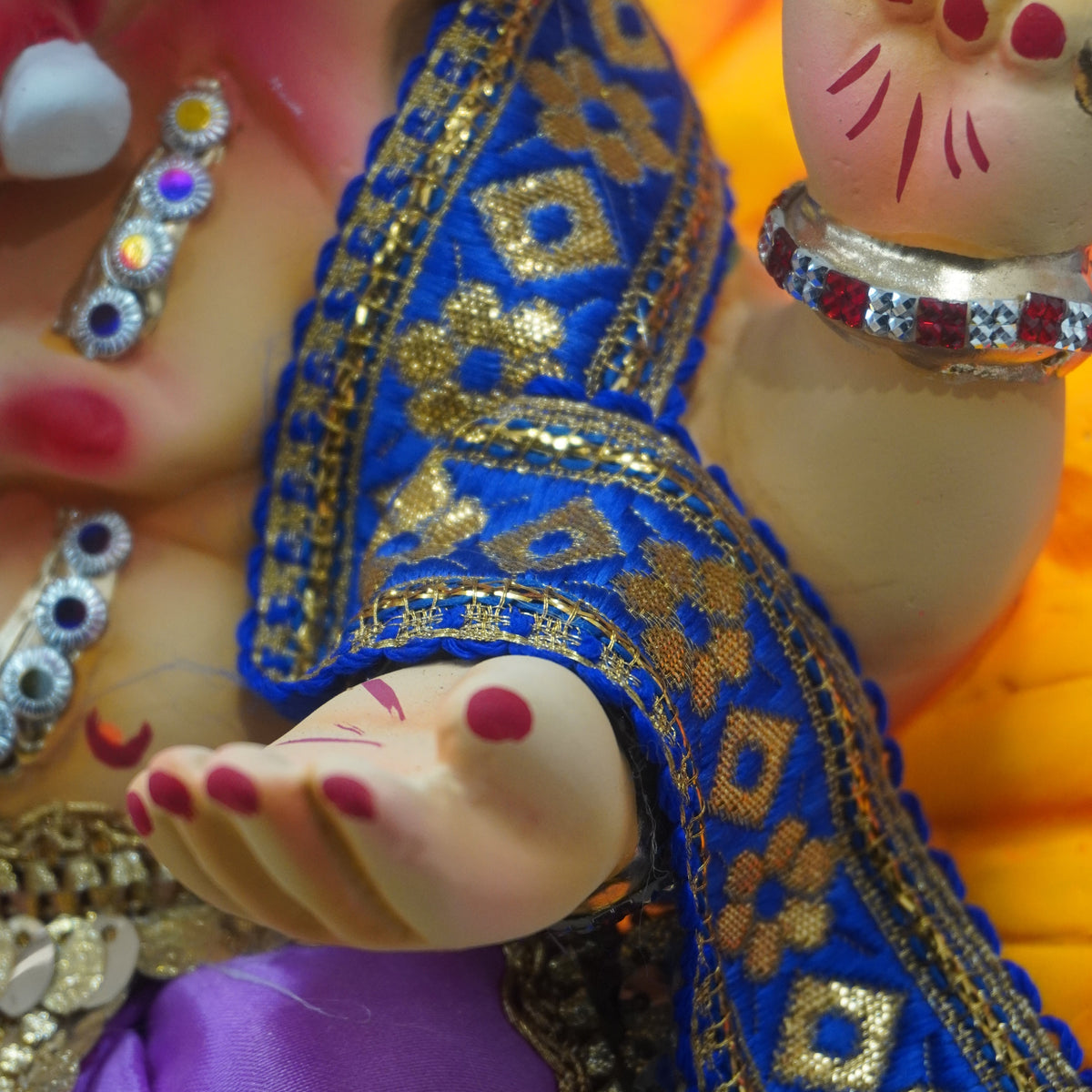 Close-up of a decorated Ganesha figurine showing the hand with red nail polish, gold jewelry, a blue embroidered sash, and a purple draped cloth.