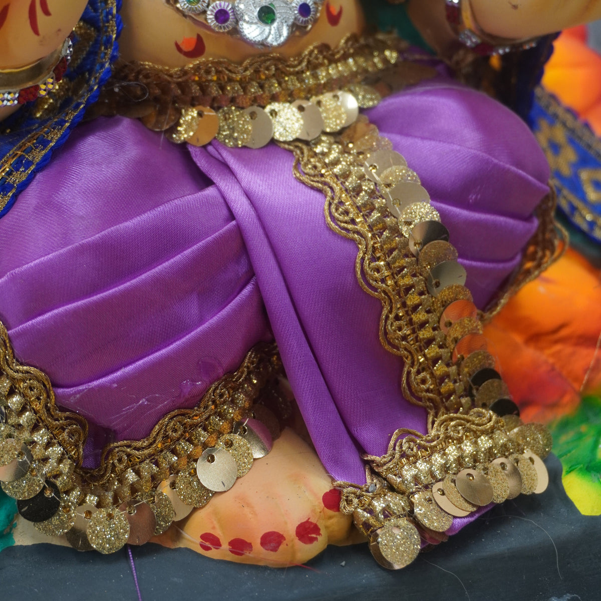 Close-up of a decorated Ganesha idol, showing the purple drape, gold coin embellishments, and colorful jewelry against a dark background.