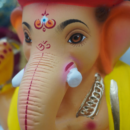Close-up of a Ganesha figurine or statue with a red crown, yellow clothing, metallic silver ornaments, and a pinkish-beige face with painted facial details.