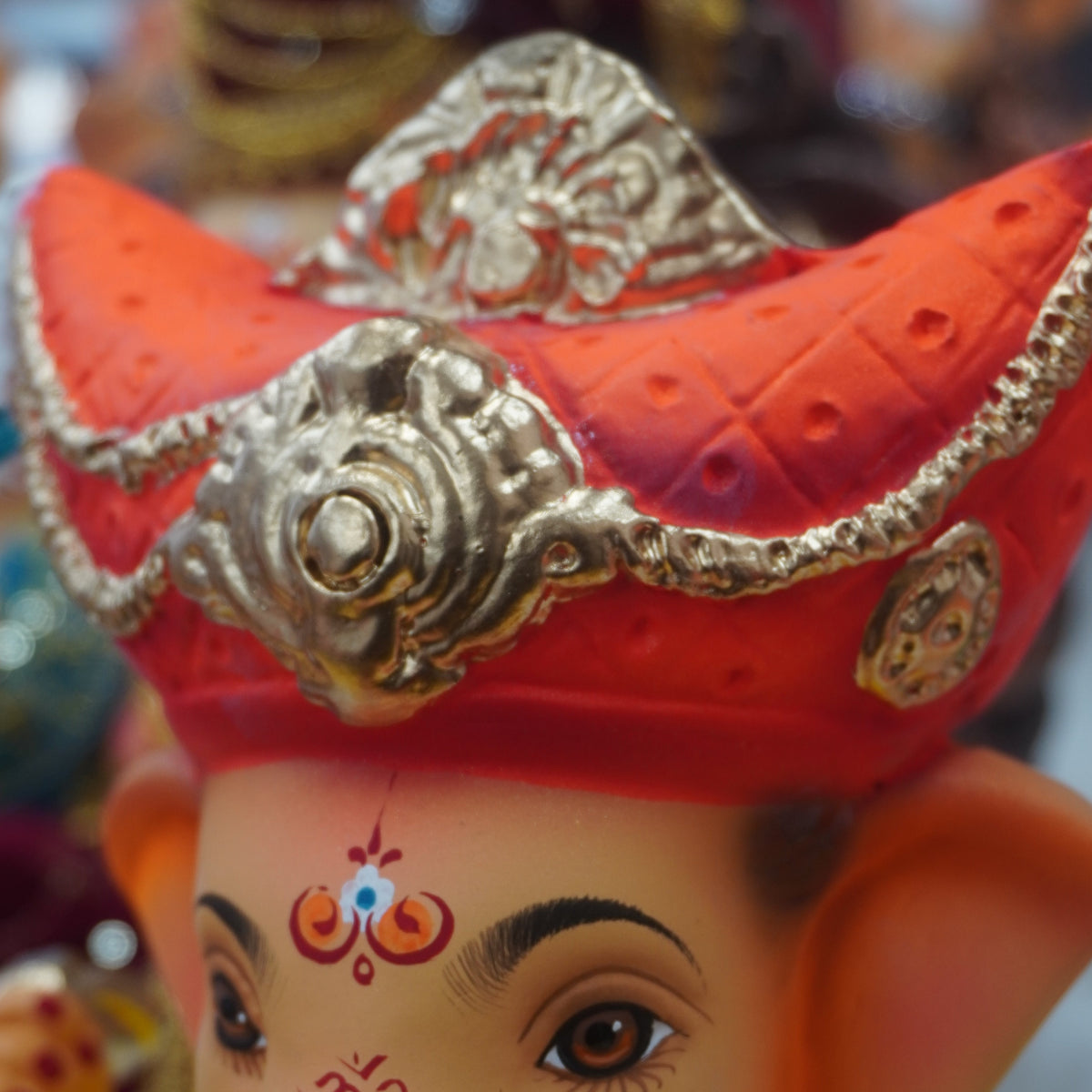 A close-up of a Ganesha idol with a bright orange-red hat, ornate silver detailing, and a yellow garment, shown against a blurred background.