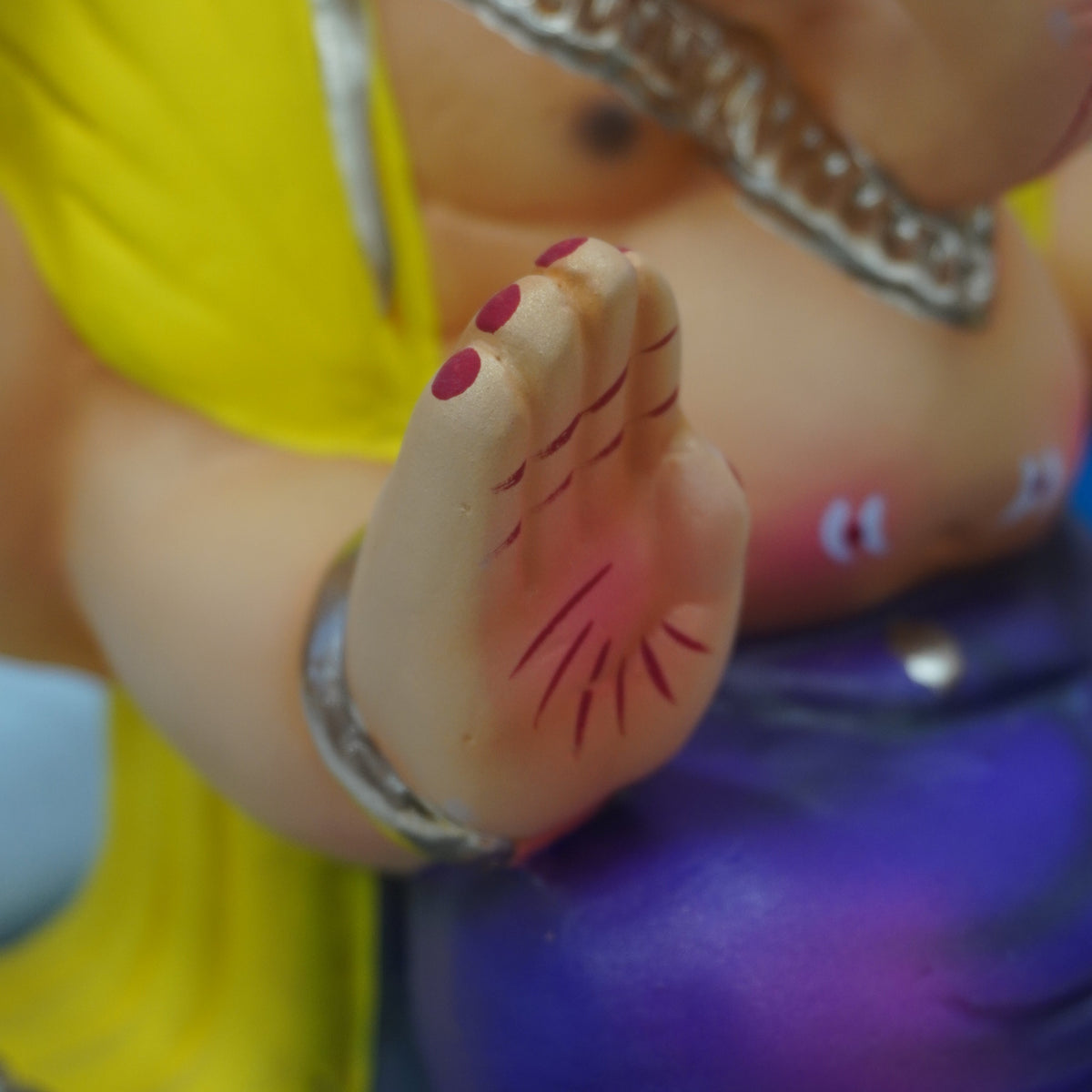 Close-up of a colorful figurine showing a human-like hand with red nail polish and red line markings, wearing a yellow garment and silver bangles, with a purple lower body.