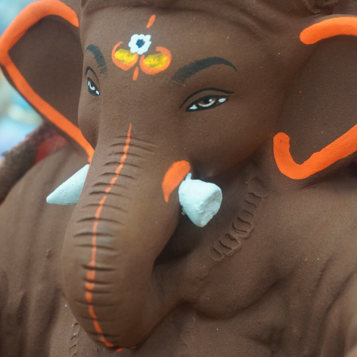 A close-up of a dark brown Ganesha statue decorated with bright orange accents, white tusks, and traditional facial paint details.