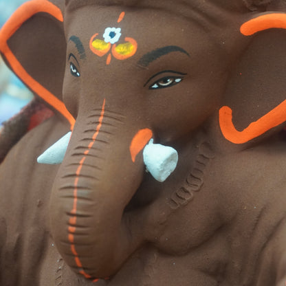 A close-up of a dark brown Ganesha statue decorated with bright orange accents, white tusks, and traditional facial paint details.