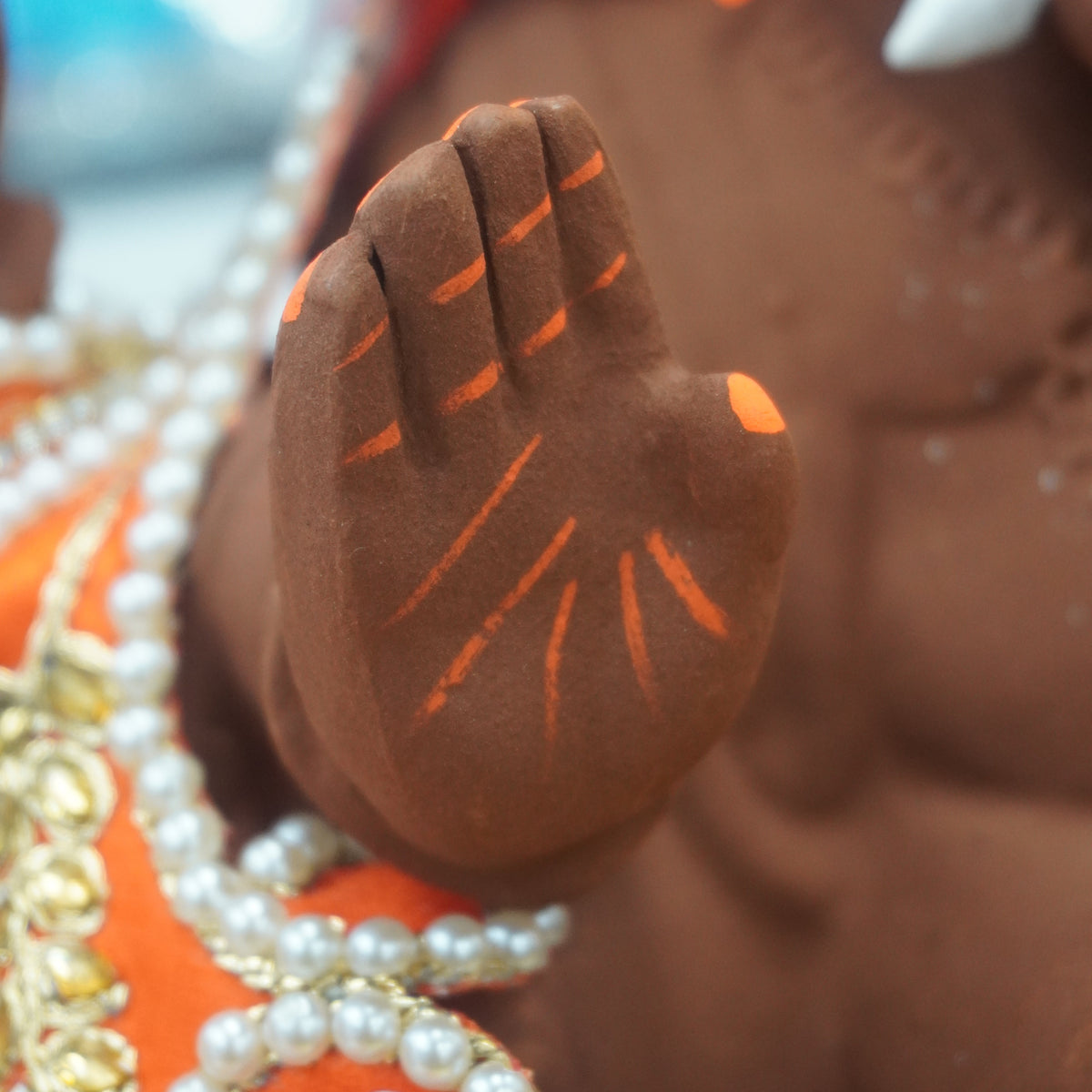 Close-up of a brown Ganesha idol hand with orange painted markings, adorned with white pearl beads and an orange fabric with a purple flower detail.