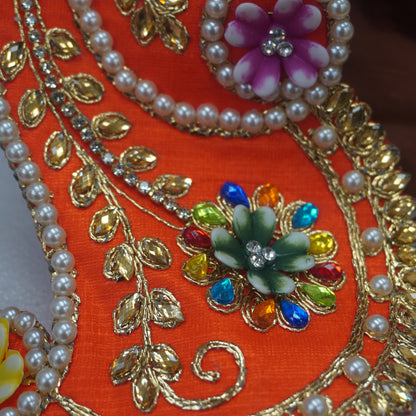 Close-up of an orange embroidered traditional garment detail with gold metallic leaf motifs, white pearl bead borders, and colorful floral embellishments in pink, green, blue, and yellow.