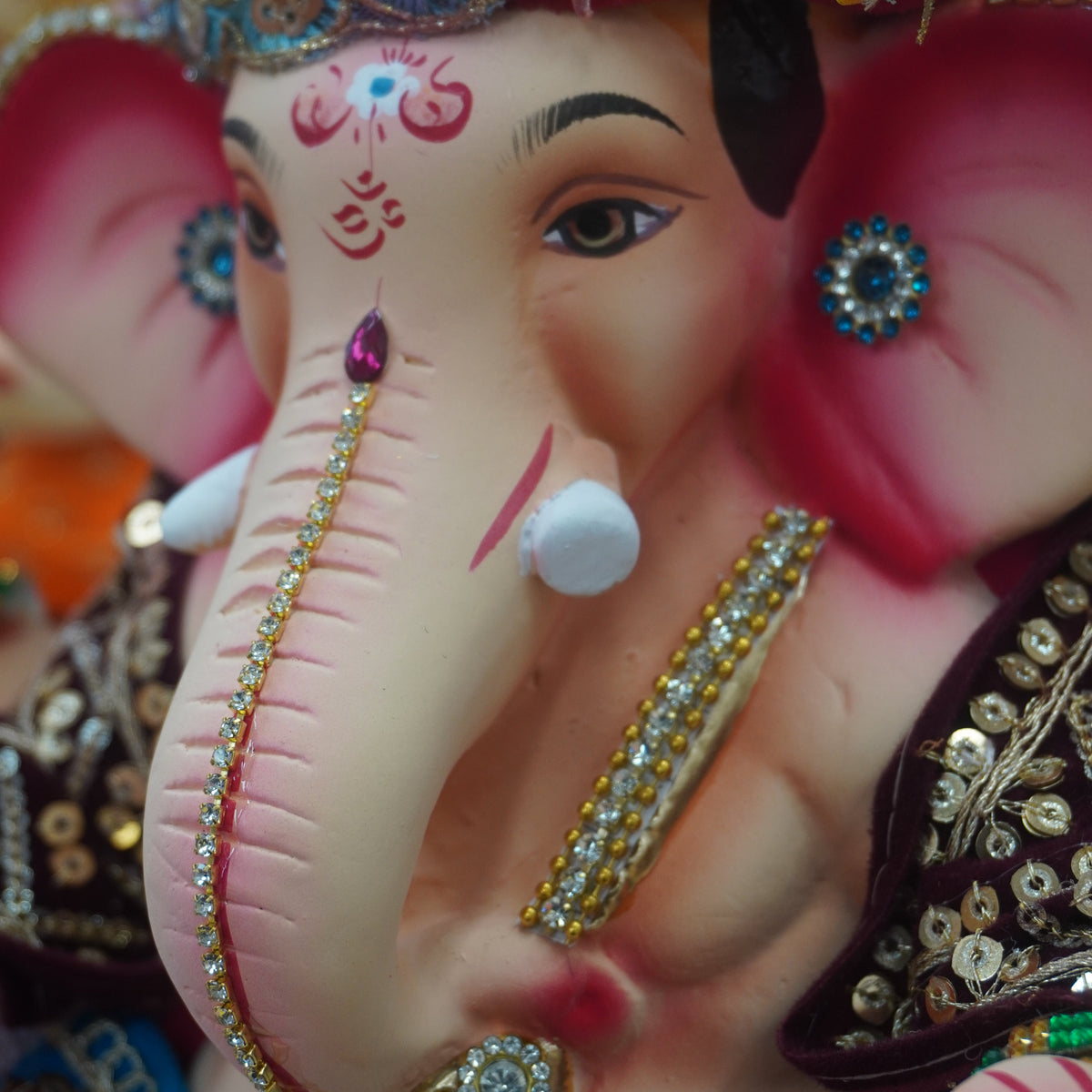 A close-up of a decorated Ganesha idol with a pink elephant head, yellow turban, and ornate gold and blue jewelry against a colorful festive background.