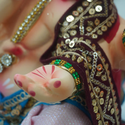Close-up of a decorative doll or figurine with a pale pink face and hand, wearing a dark embroidered sari with gold sequins and green bead accents, along with multiple gold rings.