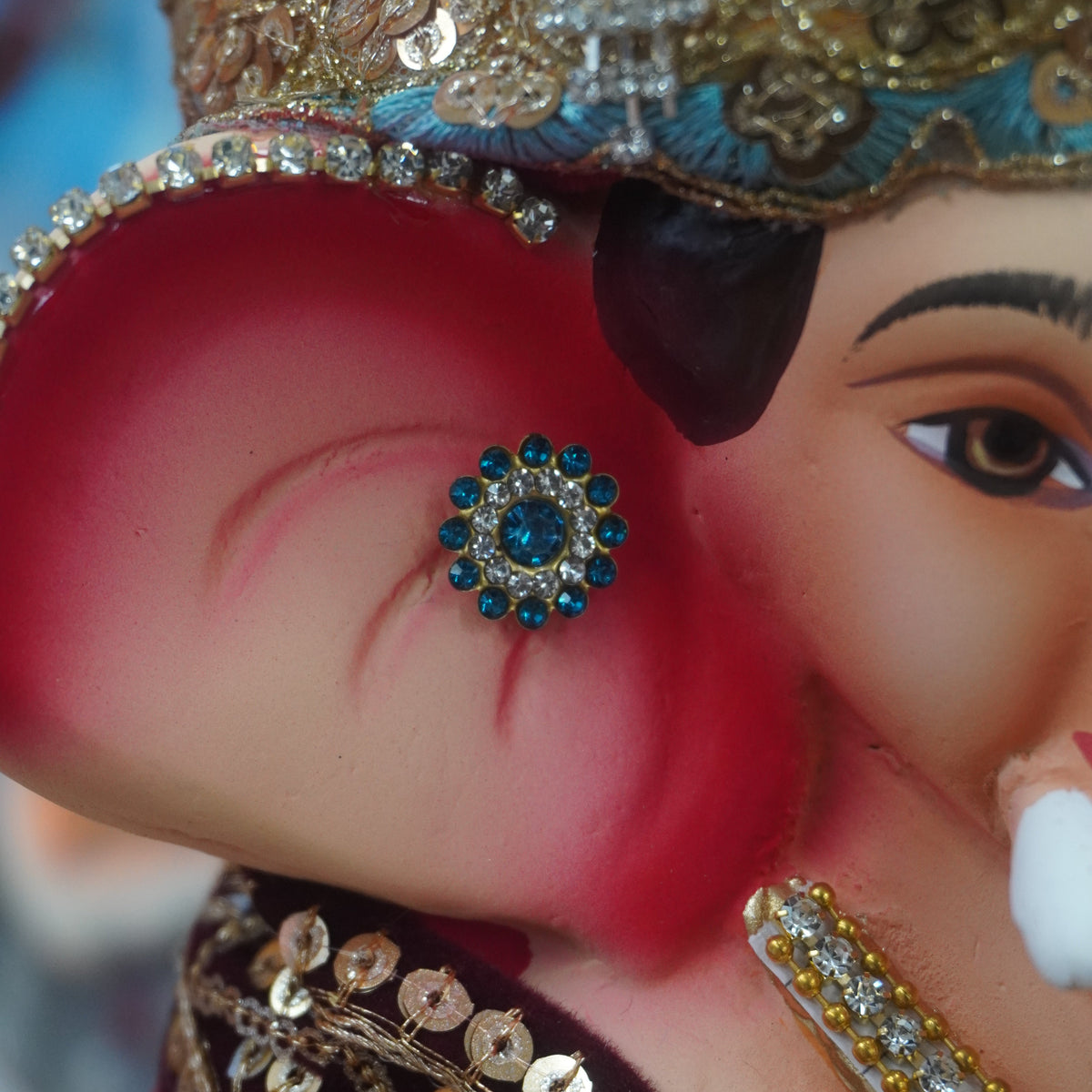 Close-up of a decorated Ganesha idol or figurine, showing the elephant ear with a blue jewel, ornate gold and silver headpiece, and embellished clothing.