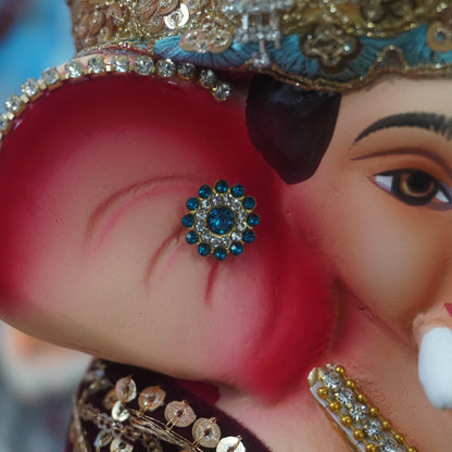 Close-up of a decorated Ganesha idol or figurine, showing the elephant ear with a blue jewel, ornate gold and silver headpiece, and embellished clothing.