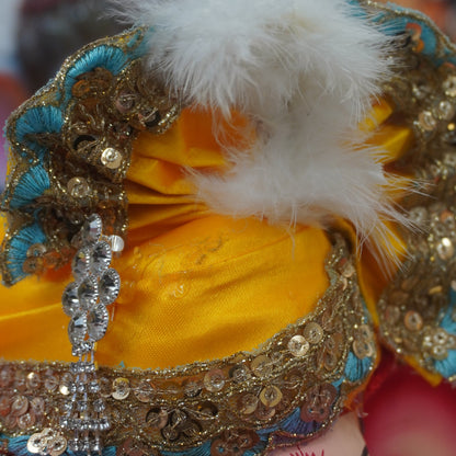 Close-up of a decorated Ganesha idol or figurine, showing the yellow turban wih golden sequin work, white feather and teal.