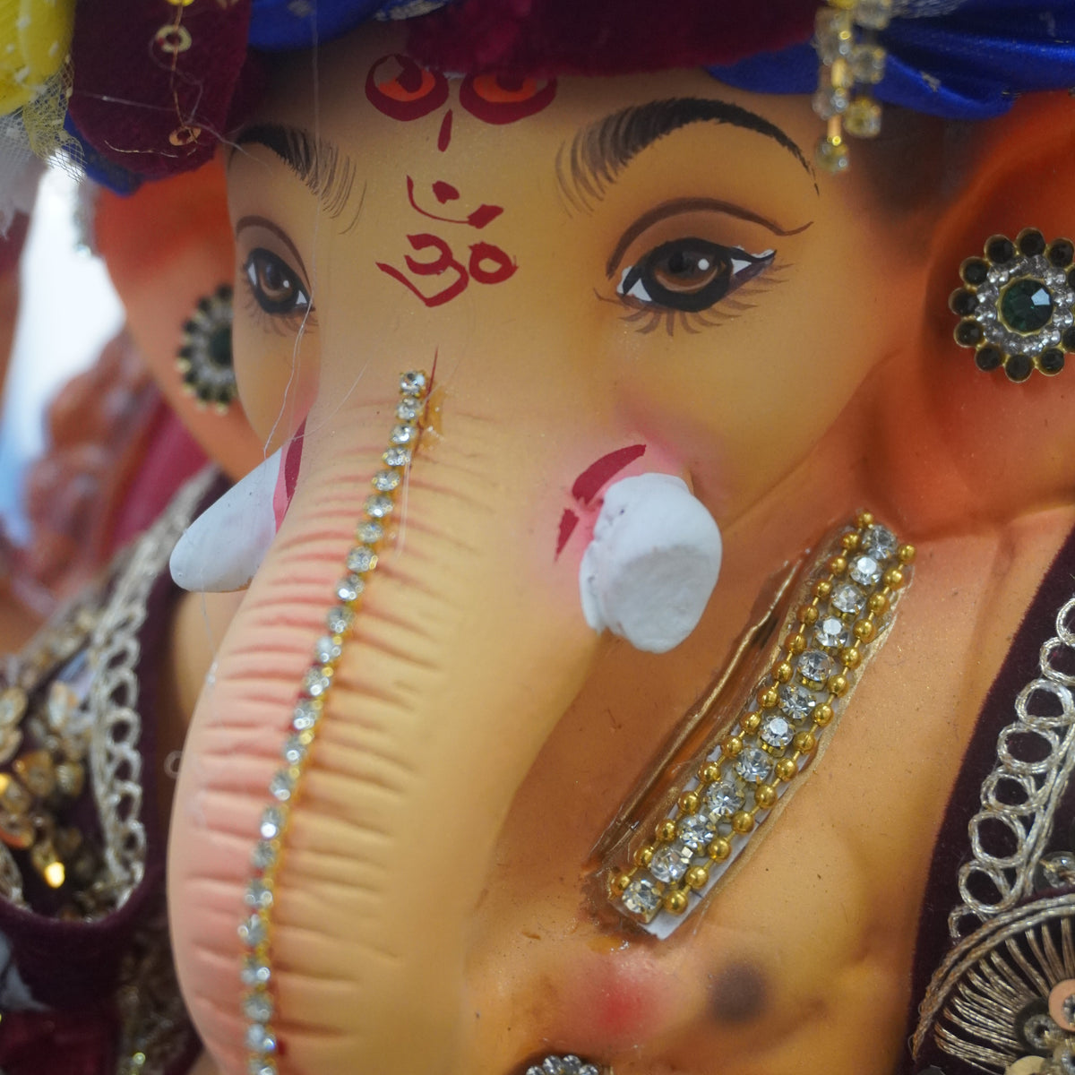 Close-up of a decorated Ganesha idol or statue, showing the elephant head, painted face, blue and maroon turban, and ornate jeweled accessories.