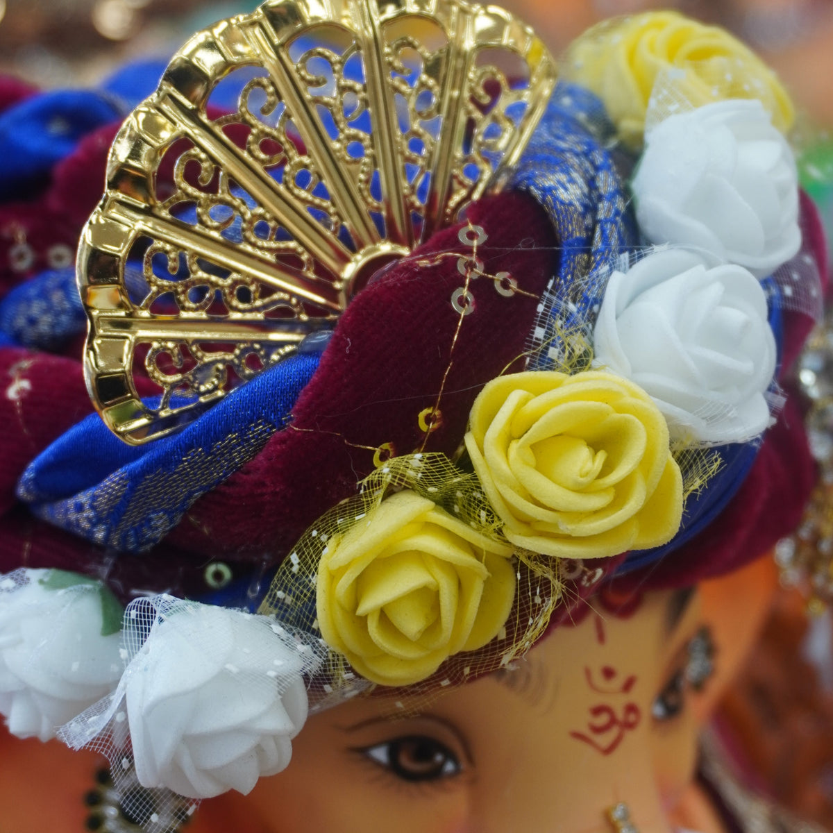 A close-up of a decorated Ganesha idol with a gold fan, blue and burgundy fabric, white and yellow flowers, and silver jewelry, shown against a blurred colorful background.