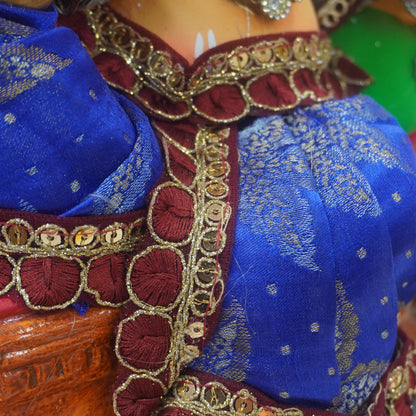 Close-up of a decorative figurine or doll wearing a royal blue sari with gold motifs and deep red scalloped borders with metallic gold trim.